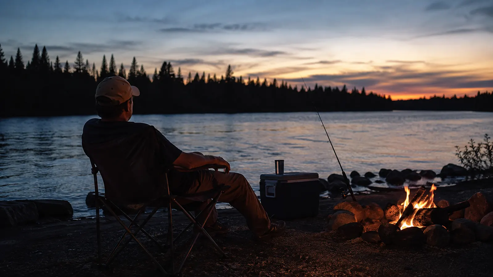 An angler leaning back in a folding camp chair by a small fire, rod propped against a cooler, dusk sky above the treeline