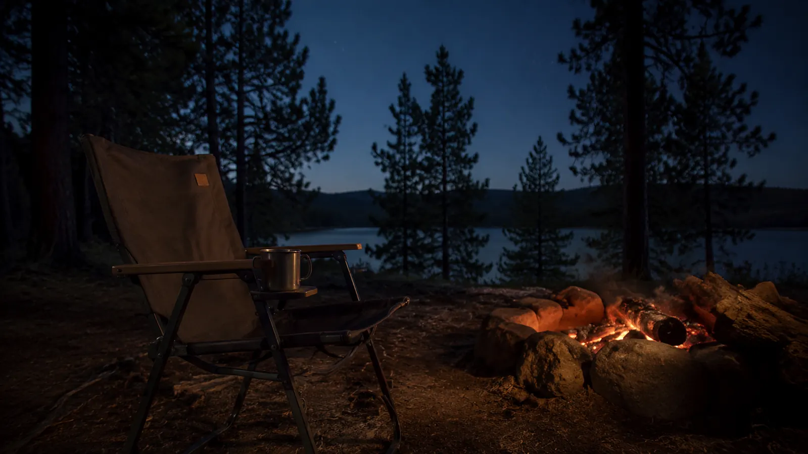 An empty camp chair angled toward a fire burned down to orange coals, a tin mug on the chair arm, dark treeline and early stars overhead.
