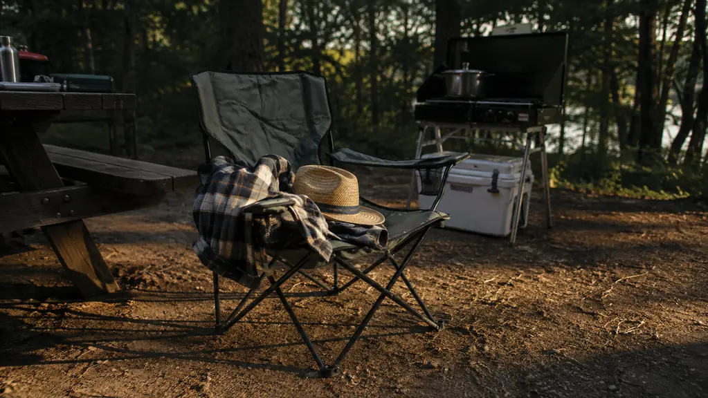 A camping chair with a wide-brim straw hat and a folded jacket on the seat at a shaded campsite in late afternoon, a camp stove and cooler nearby