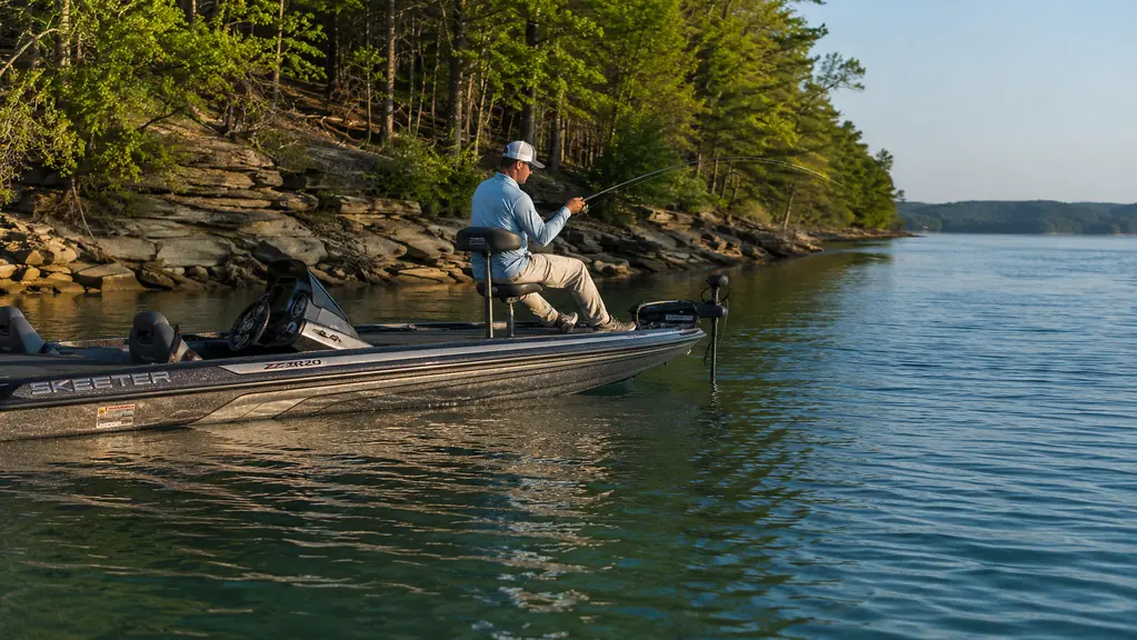 A bass boat anchored over a shadowed drop-off in clear water, late afternoon light on a tree-lined bank