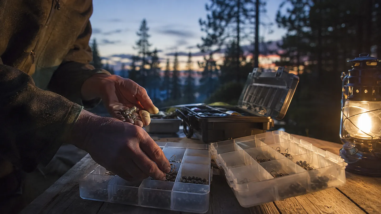 An angler sorting hooks and split-shot weights from an open tackle box into empty divided organizer trays on a camp table at dusk, a lantern glowing, pines behind